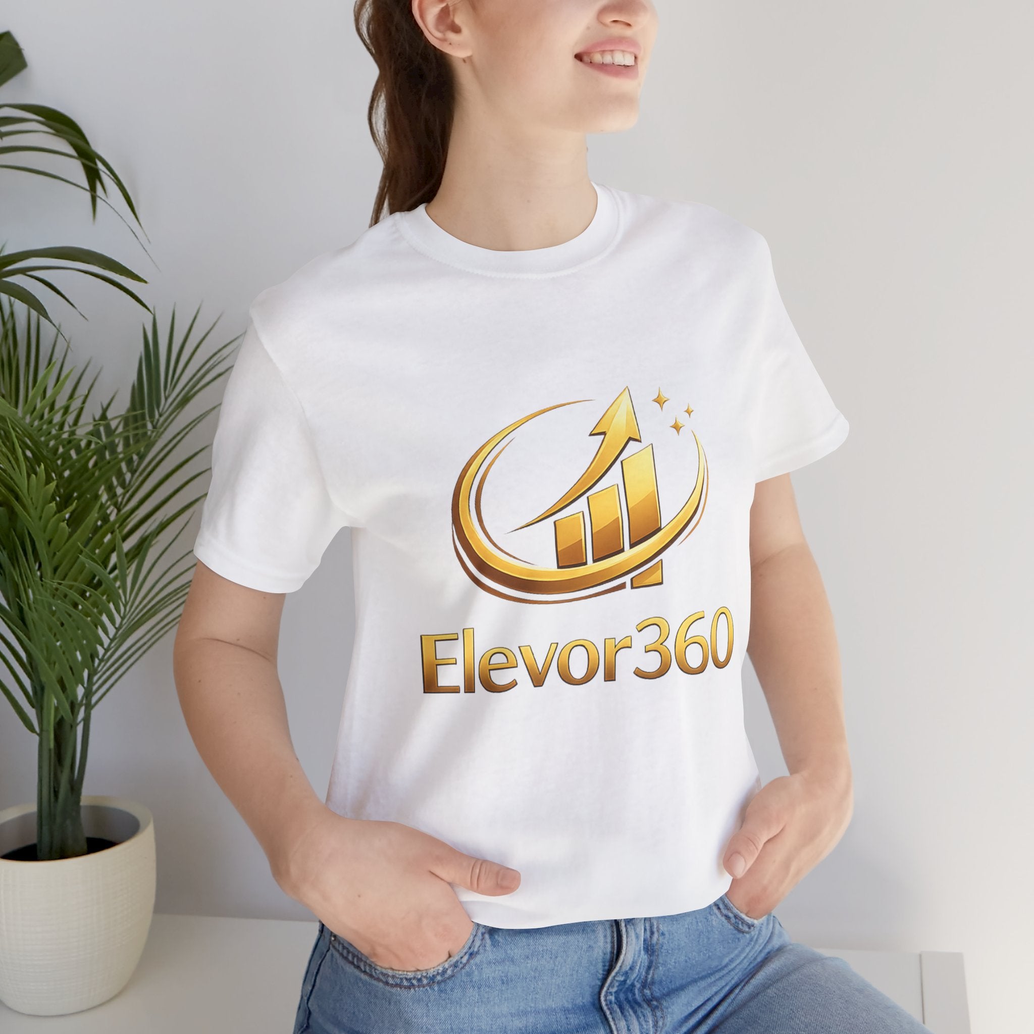 Person wearing a white t-shirt with 'Elevor360' logo in gold, standing indoors next to a plant.