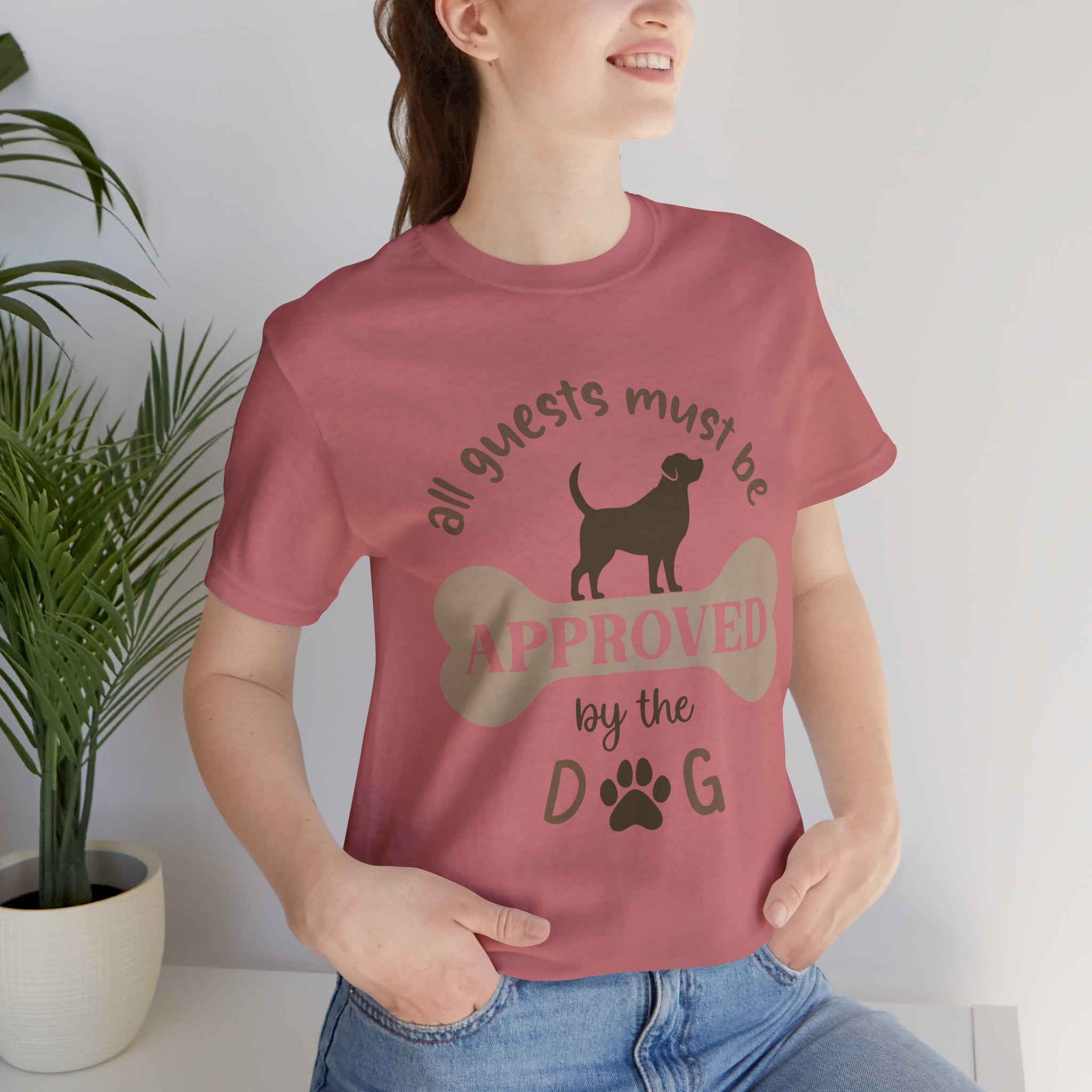 Person wearing a pink t-shirt with a dog-themed design, standing indoors next to a plant.