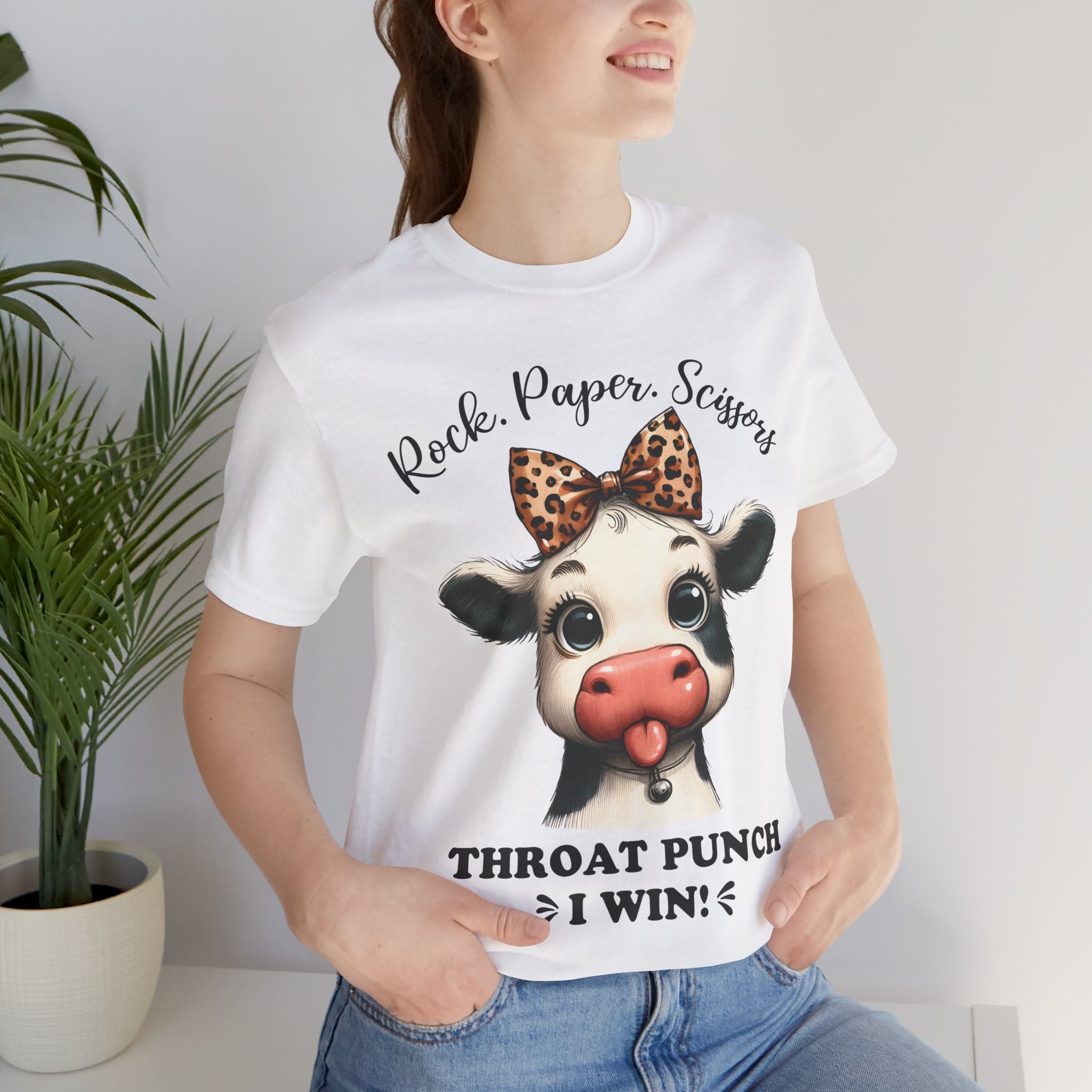 Person wearing a white t-shirt with a cow graphic and text, standing indoors next to a plant.